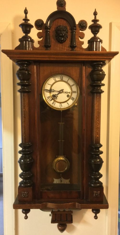 fig 1. A dark walnut Vienna regulator wall clock featuring carved finials, side pilasters, a white enamel dial with Roman numerals, and a brass pendulum behind a glass door.