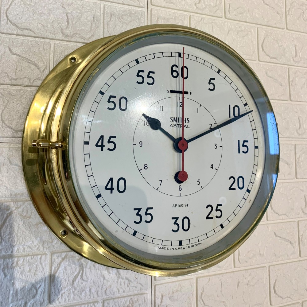 A circular Smiths Astral brass marine clock featuring a white dial face, black Arabic numerals, and a red centre hand mounted against a wall.
