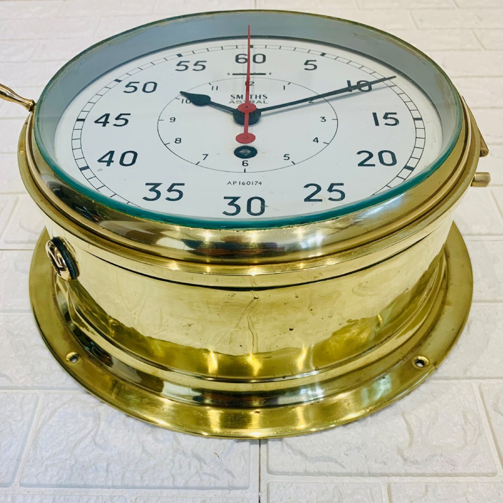 A circular Smiths Astral brass marine clock featuring a white dial face, black Arabic numerals, and a red centre hand mounted against a wall. View II