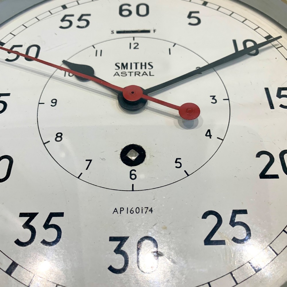 A circular Smiths Astral brass marine clock featuring a white dial face, black Arabic numerals, and a red centre hand mounted against a wall. View III