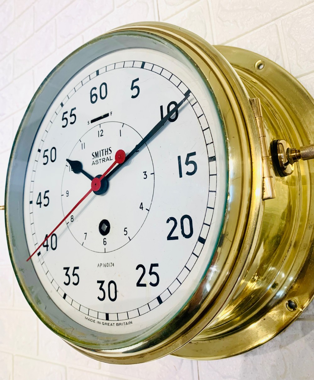 A circular Smiths Astral brass marine clock featuring a white dial face, black Arabic numerals, and a red centre hand mounted against a wall. View IV
