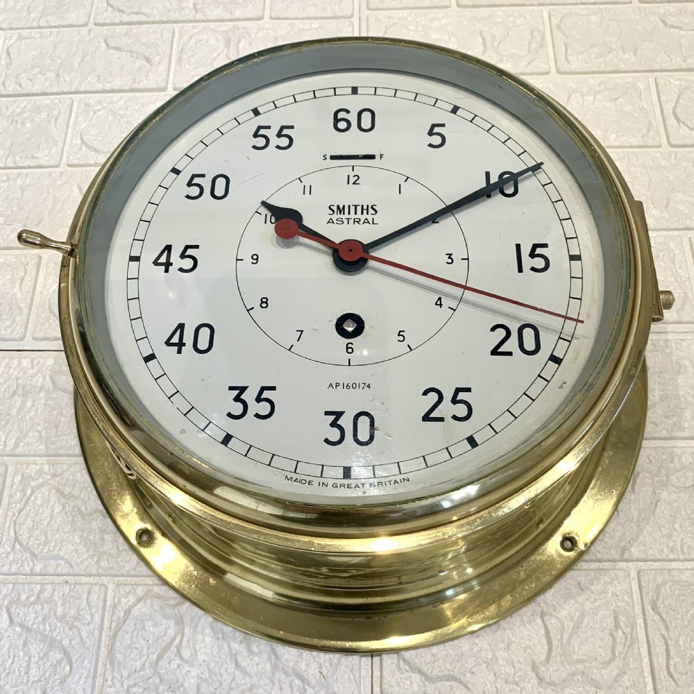 A circular Smiths Astral brass marine clock featuring a white dial face, black Arabic numerals, and a red centre hand mounted against a wall. View VI