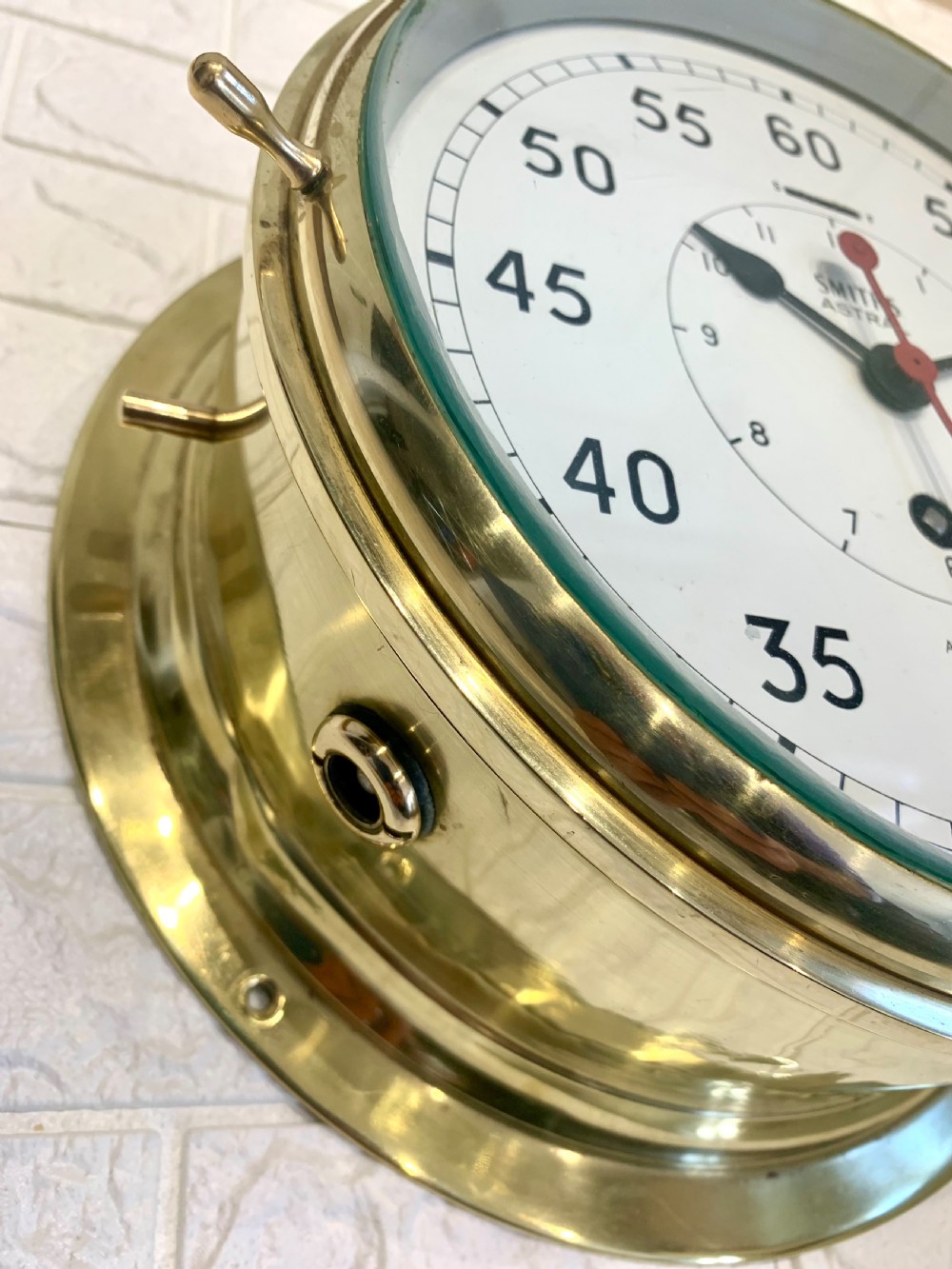 A circular Smiths Astral brass marine clock featuring a white dial face, black Arabic numerals, and a red centre hand mounted against a wall. View VII