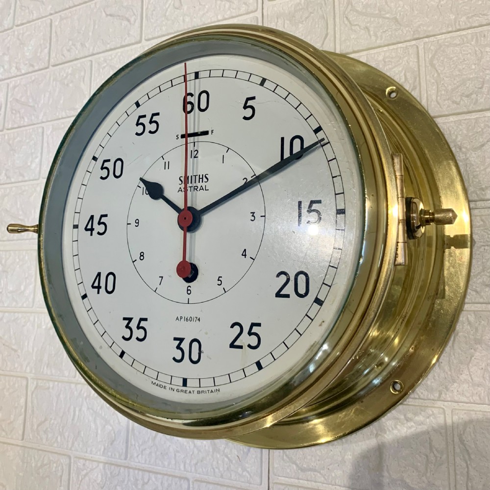 A circular Smiths Astral brass marine clock featuring a white dial face, black Arabic numerals, and a red centre hand mounted against a wall. View VIII