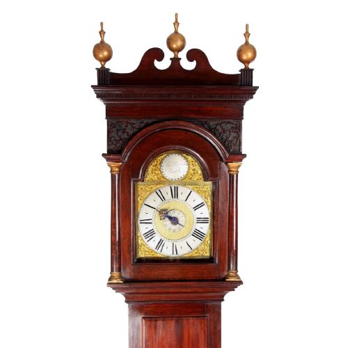 fig 1. A Georgian mahogany longcase clock featuring three brass finials, an arched brass dial with ornate spandrels, and a tall slender wooden case.