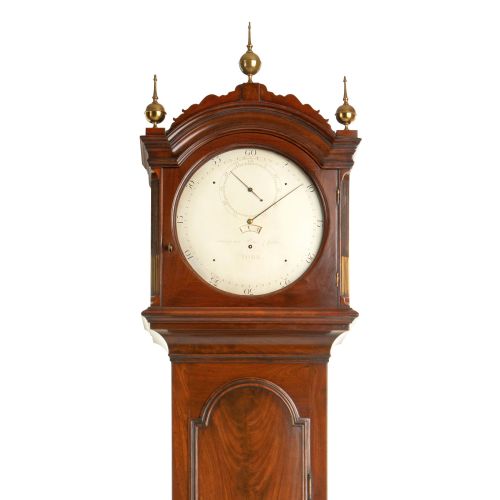 fig 1. An antique mahogany longcase regulator clock with three brass finials on an arched hood, a white regulator dial, and a flame mahogany trunk door.