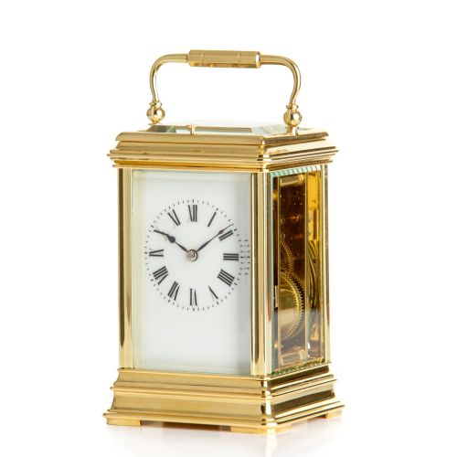 fig 1. A rectangular polished brass French carriage clock featuring a top carrying handle, bevelled glass side panels, and a white enamel dial with Roman numerals.
