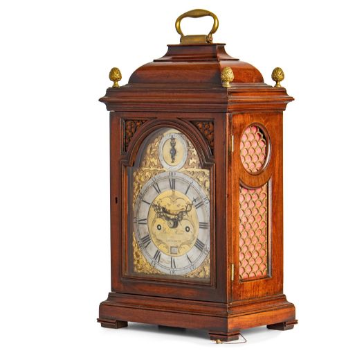 fig 1. A Georgian mahogany striking table clock featuring an arched brass and silvered dial, decorative corner spandrels, a bell top with brass handle, and pineapple finials.