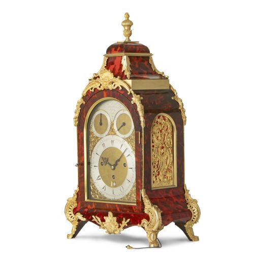 fig 1. An arched red tortoiseshell bracket clock adorned with gilded ormolu mounts, Rococo scroll feet, and a silvered dial.