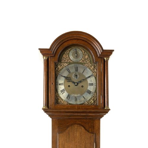 fig 2. A compact oak longcase clock featuring an arched hood, brass dial with Roman numerals, and a waist door with a hanging tassel.
