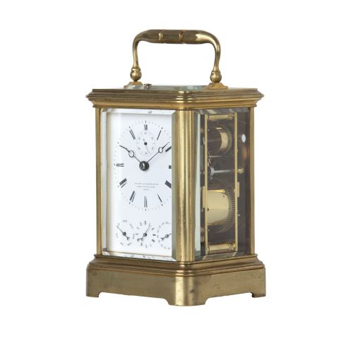 fig 3. A rectangular brass carriage clock featuring a top carrying handle, glass panels, a main white enamel dial with Roman numerals, and three subsidiary dials for calendar functions.
