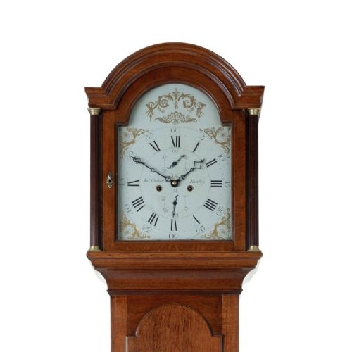 fig 2. An antique oak longcase clock featuring a rounded arched hood, a white painted dial with Roman numerals, and a tall slender trunk set on a plinth base.