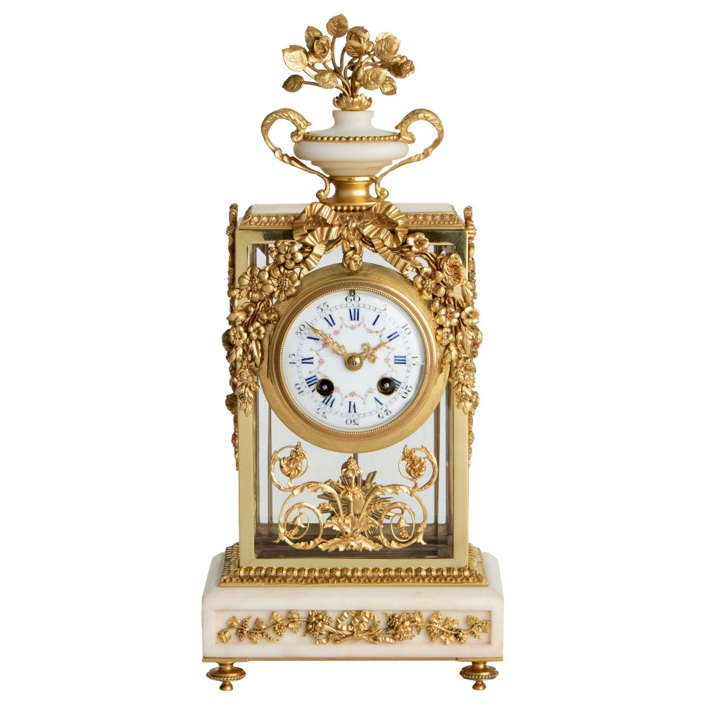 A French Neoclassical gilt bronze and white marble mantel clock featuring a glass case, an enamelled dial, and an urn finial decorated with floral motifs.