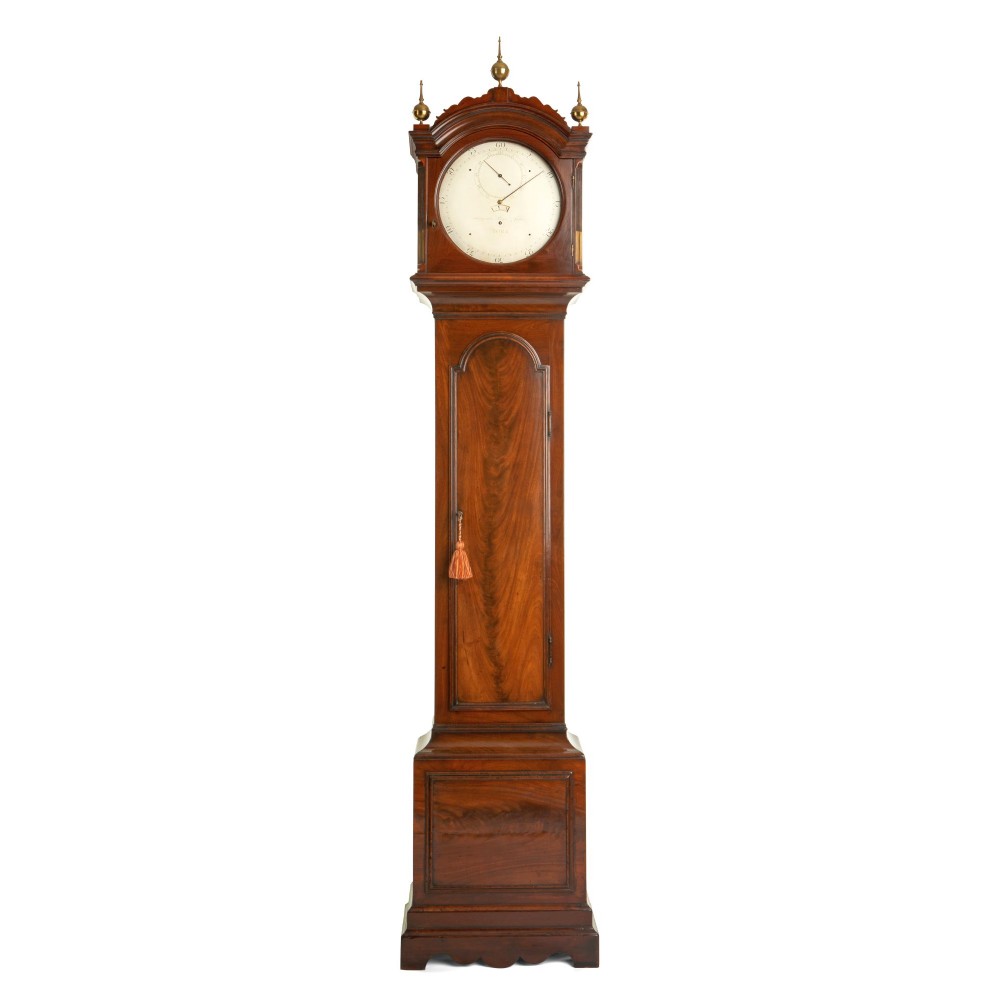 An antique mahogany longcase regulator clock with three brass finials on an arched hood, a white regulator dial, and a flame mahogany trunk door.
