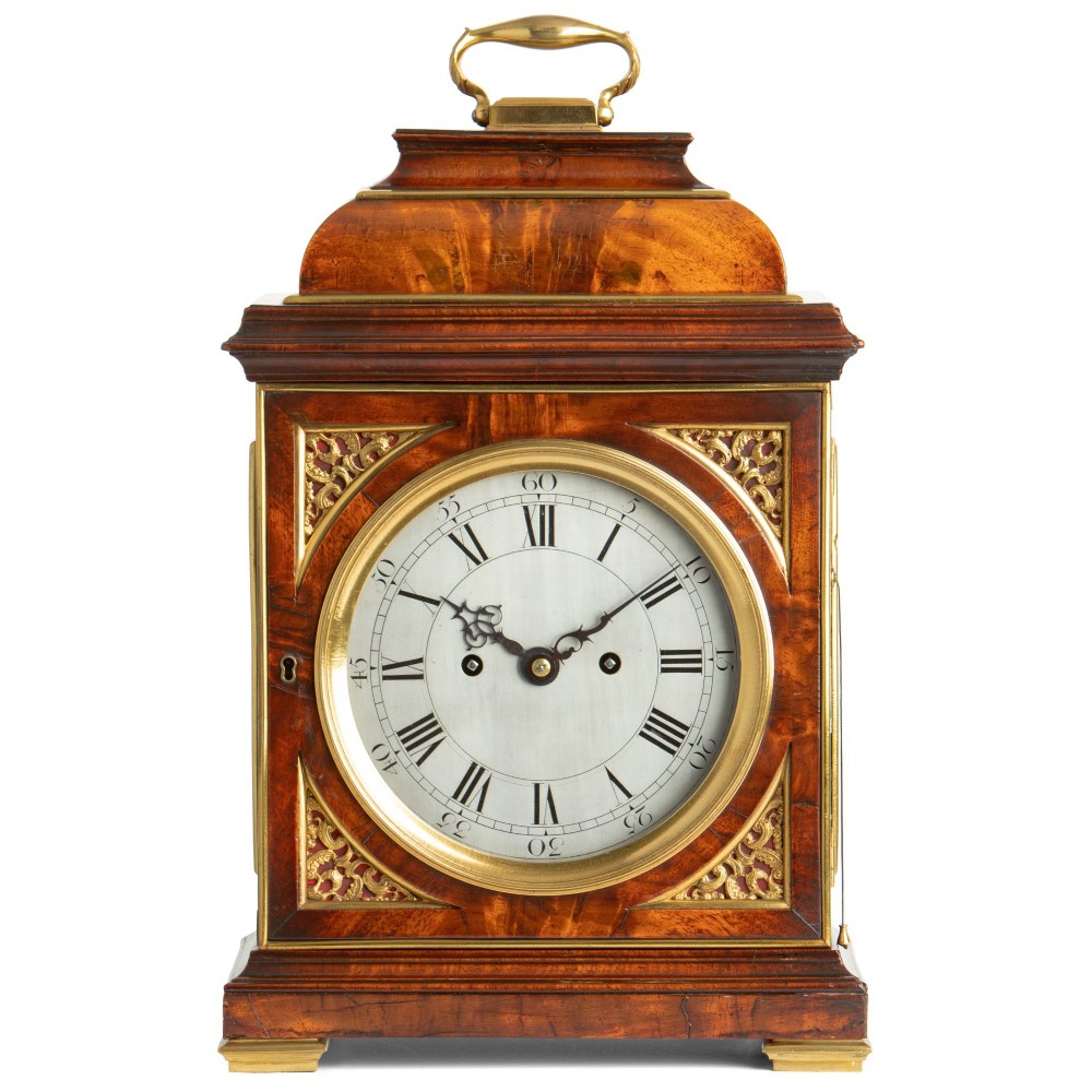 A Georgian mahogany table clock featuring an arched bell top with a brass carrying handle, gilt brass spandrels, and a circular silvered dial with Roman numerals.