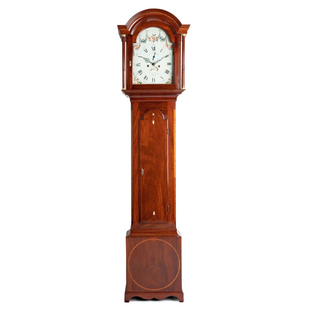 An antique mahogany Channel Island longcase clock featuring a painted arched dial with floral detail, an inlaid waist door, and turned hood columns.