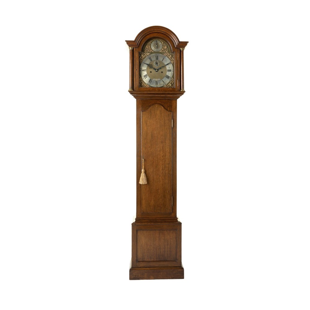 A compact oak longcase clock featuring an arched hood, brass dial with Roman numerals, and a waist door with a hanging tassel.