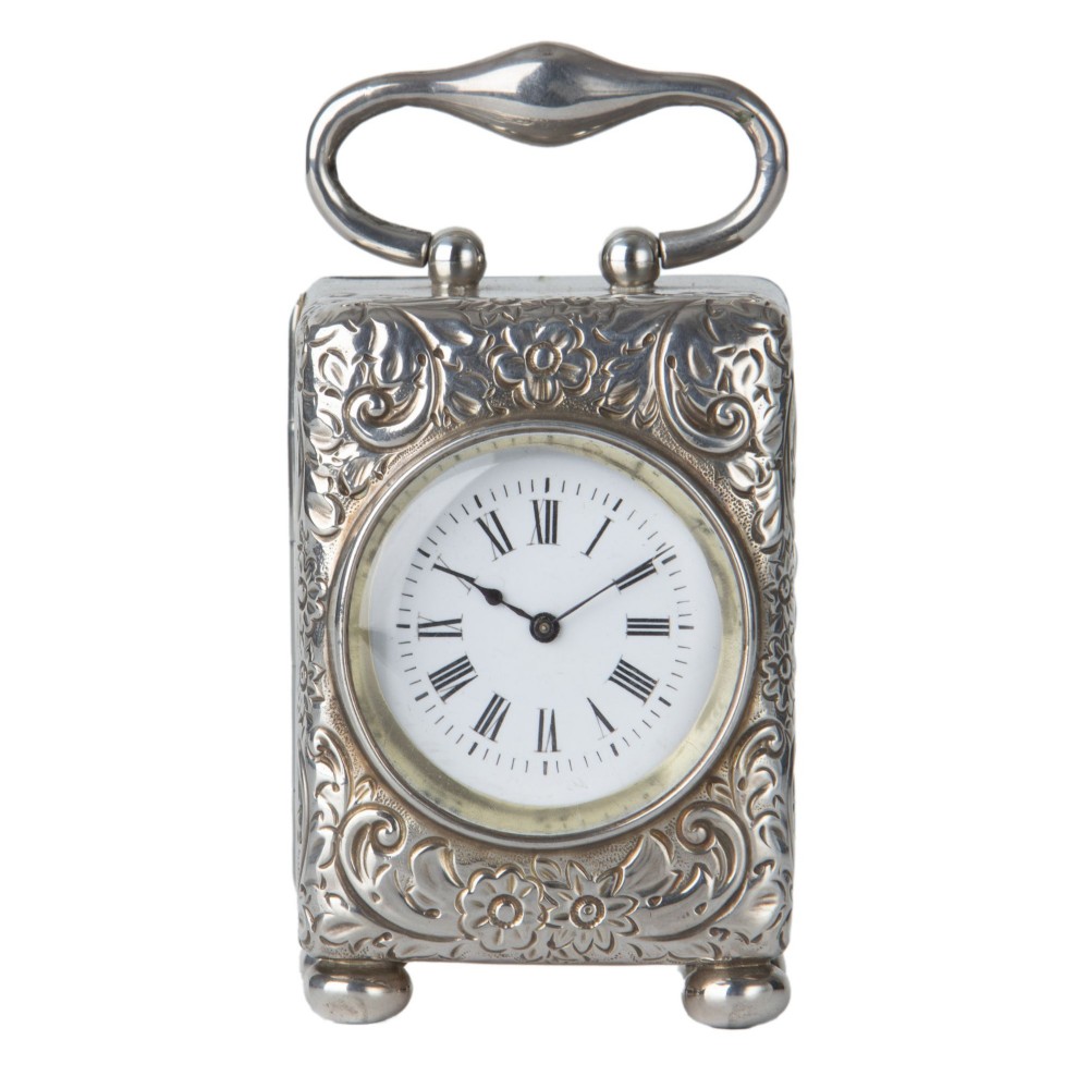 A rectangular chased silver carriage clock featuring floral scrollwork, a looped top handle, ball feet, and a white enamel dial with Roman numerals.