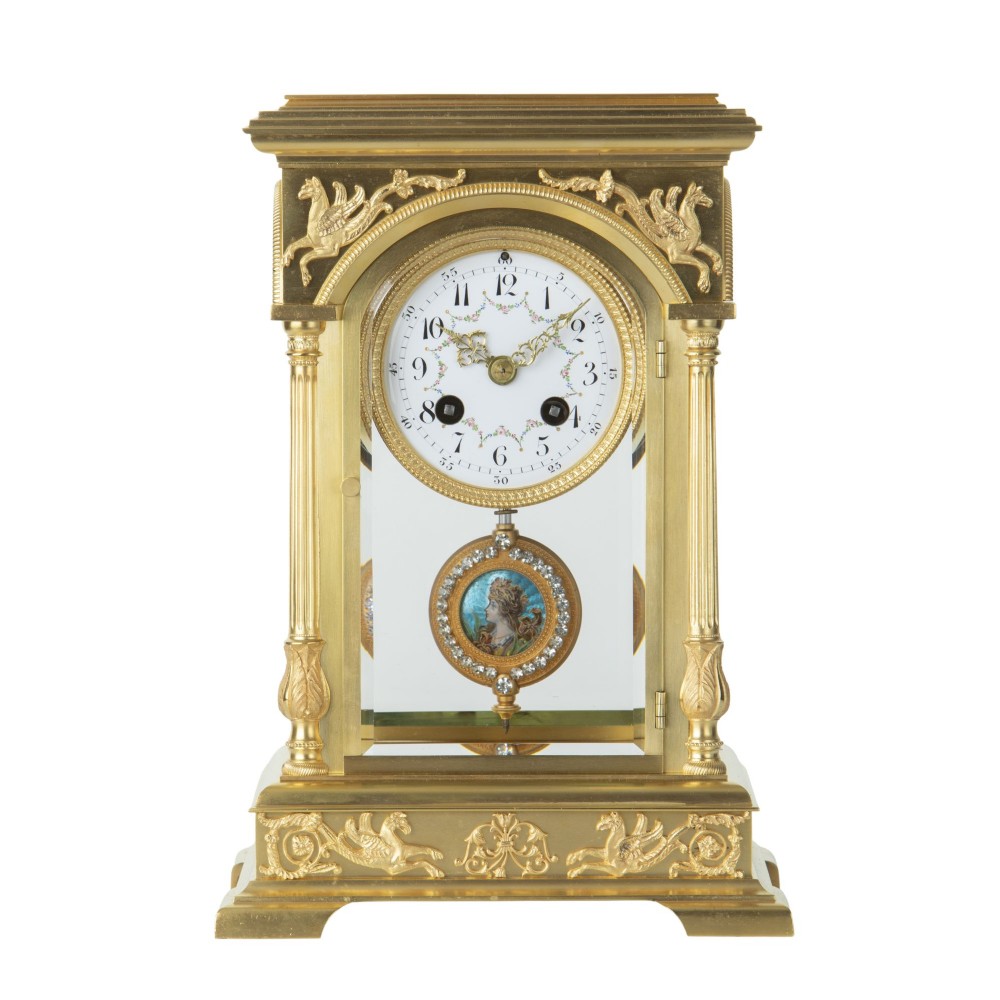 An antique gilded brass four glass mantel clock featuring fluted corner columns, winged pegasus reliefs, an enamel dial, and a decorative pendulum with a portrait miniature.