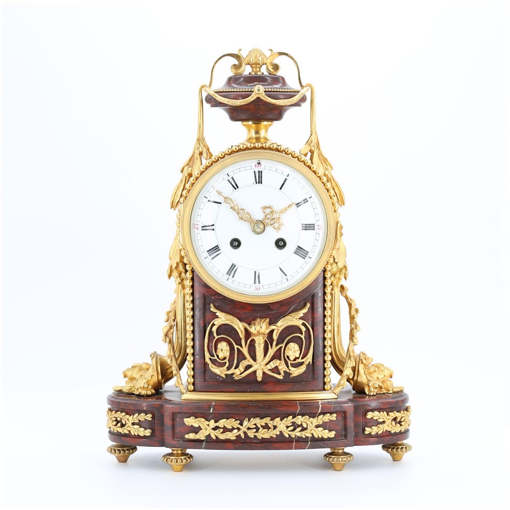 A French Louis XVI style mantel clock featuring a red marble body, gilt bronze floral garlands and urn finial, and a white enamel clock face with Roman numerals.