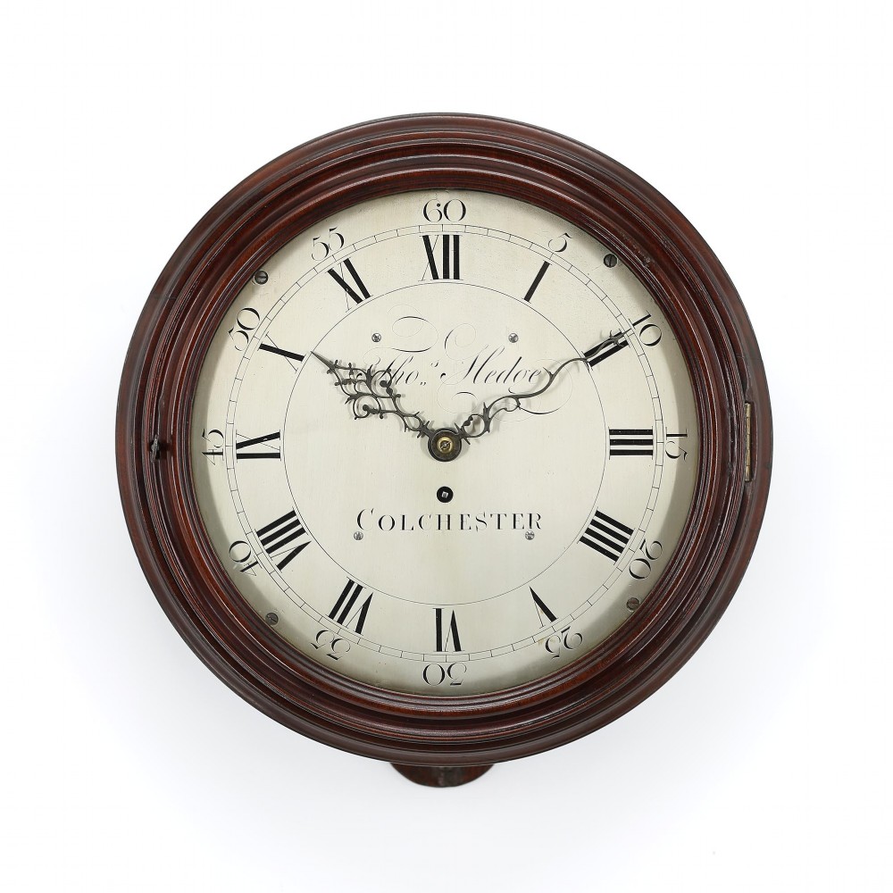 An antique circular Georgian verge dial clock featuring a moulded mahogany surround, cream painted dial with Roman numerals, and decorative black hands.