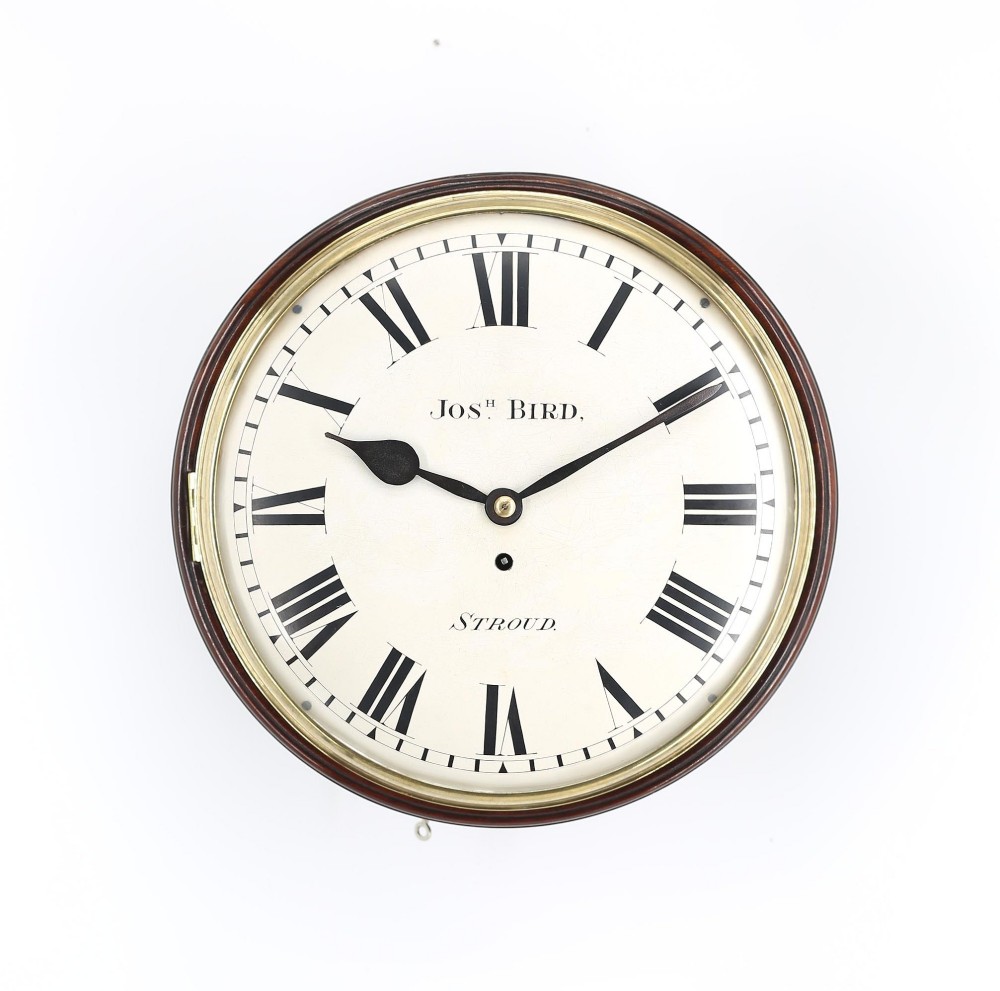 A circular antique mahogany wall clock featuring a convex off-white dial with Roman numerals, black spade hands, and a brass bezel surround.