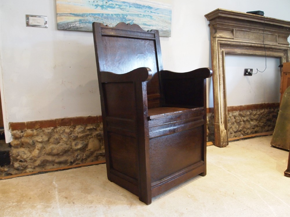 An early 18th-century dark oak lambing chair featuring a high panelled back, enclosed box base, integrated shaped arms, and a cut crest rail.