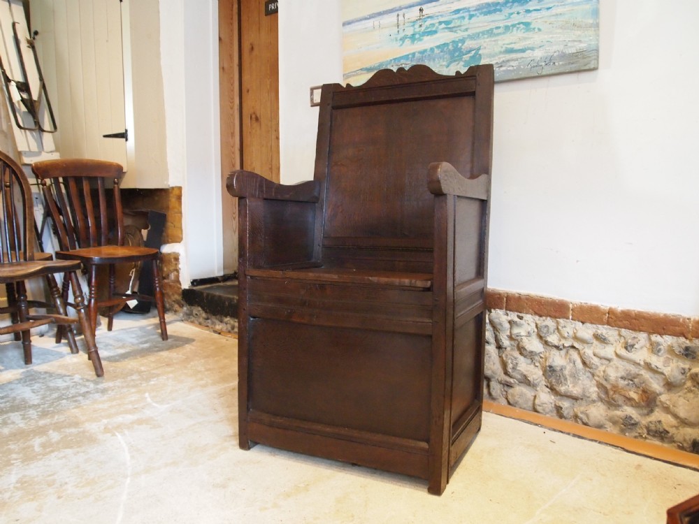 An early 18th-century dark oak lambing chair featuring a high panelled back, enclosed box base, integrated shaped arms, and a cut crest rail. View II