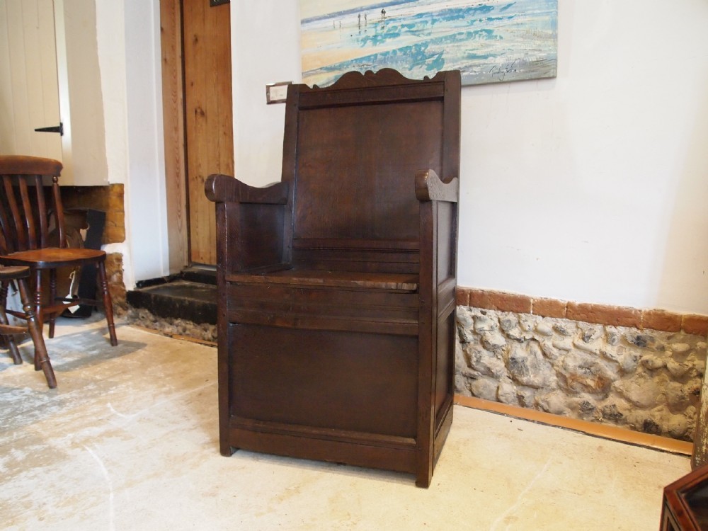 An early 18th-century dark oak lambing chair featuring a high panelled back, enclosed box base, integrated shaped arms, and a cut crest rail. View VIII