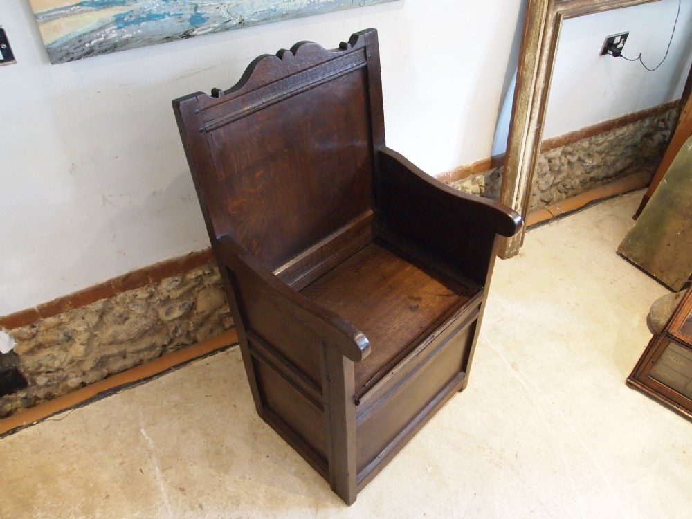 An early 18th-century dark oak lambing chair featuring a high panelled back, enclosed box base, integrated shaped arms, and a cut crest rail. View IX