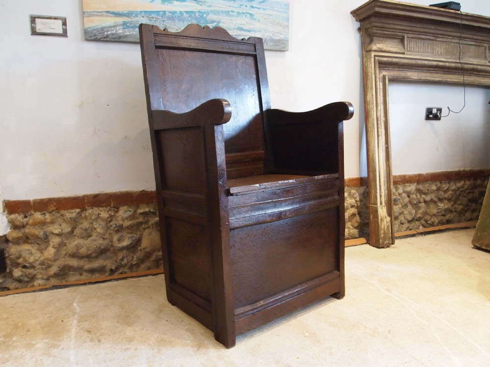 An early 18th-century dark oak lambing chair featuring a high panelled back, enclosed box base, integrated shaped arms, and a cut crest rail. View X