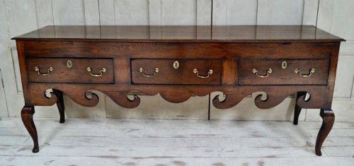 fig 1. An early Georgian dark oak dresser base featuring three drawers with brass handles, a distinctive scrolled apron, and cabriole legs.