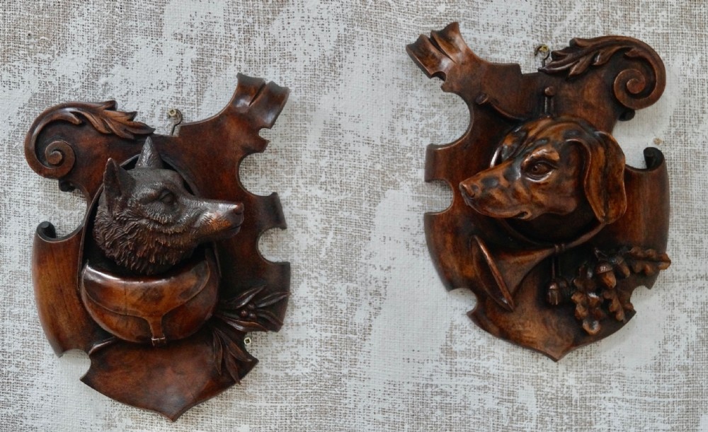 A pair of dark carved wooden shield-shaped wall plaques depicting relief heads of a fox and a hunting hound alongside sporting accessories. View X