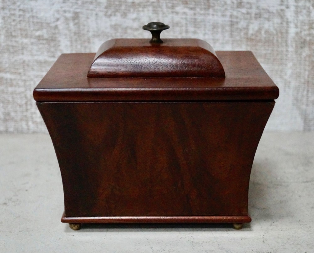A mahogany sarcophagus-shaped tea caddy featuring flared sides, a stepped hinged lid with a central brass finial, and small brass feet.