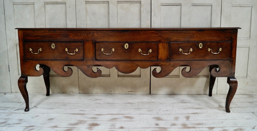 An early Georgian dark oak dresser base featuring three drawers with brass handles, a distinctive scrolled apron, and cabriole legs.