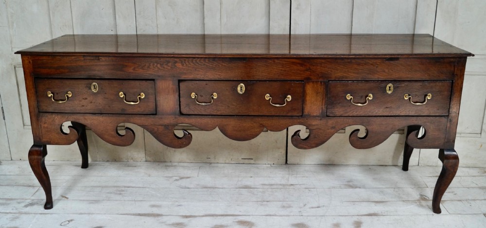 An early Georgian dark oak dresser base featuring three drawers with brass handles, a distinctive scrolled apron, and cabriole legs. View II