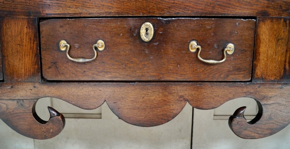 An early Georgian dark oak dresser base featuring three drawers with brass handles, a distinctive scrolled apron, and cabriole legs. View IV