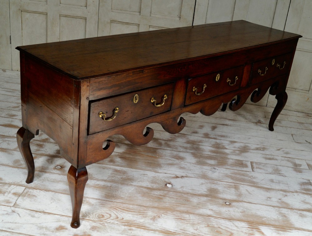 An early Georgian dark oak dresser base featuring three drawers with brass handles, a distinctive scrolled apron, and cabriole legs. View V