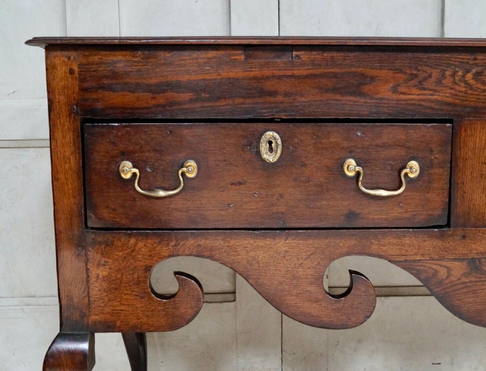 An early Georgian dark oak dresser base featuring three drawers with brass handles, a distinctive scrolled apron, and cabriole legs. View VII