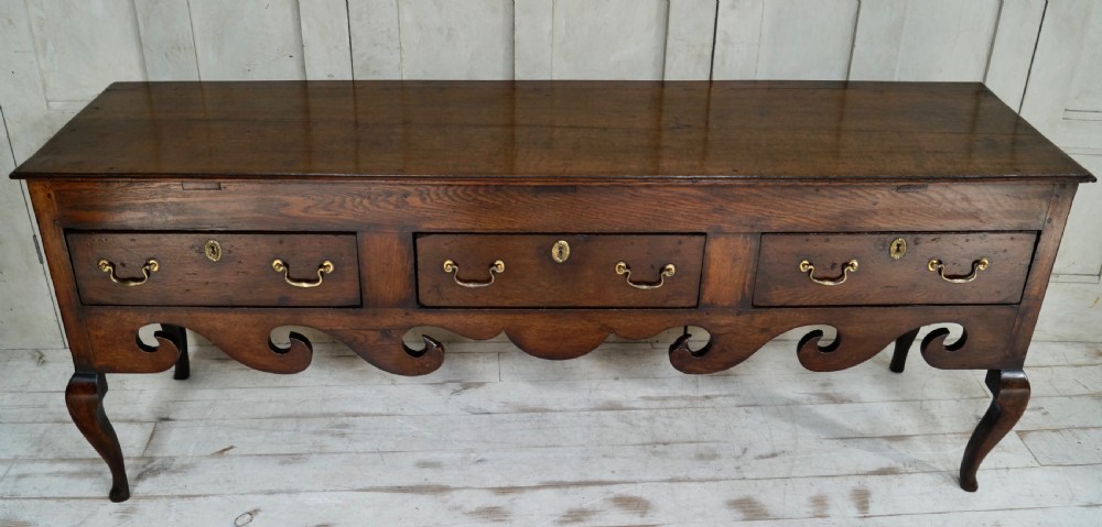 An early Georgian dark oak dresser base featuring three drawers with brass handles, a distinctive scrolled apron, and cabriole legs. View VIII