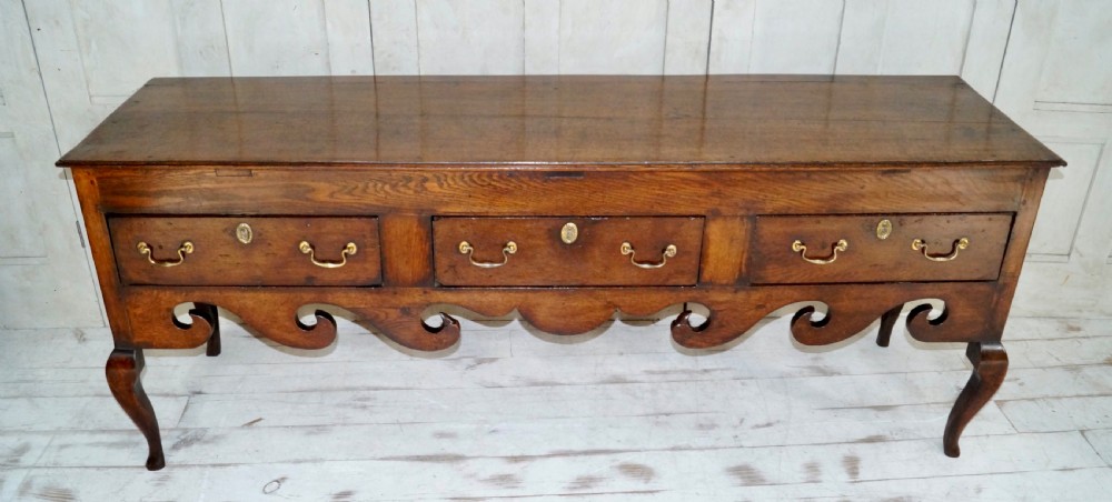 An early Georgian dark oak dresser base featuring three drawers with brass handles, a distinctive scrolled apron, and cabriole legs. View IX