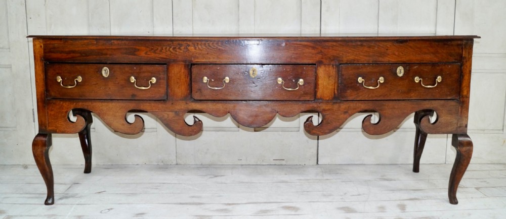 An early Georgian dark oak dresser base featuring three drawers with brass handles, a distinctive scrolled apron, and cabriole legs. View X