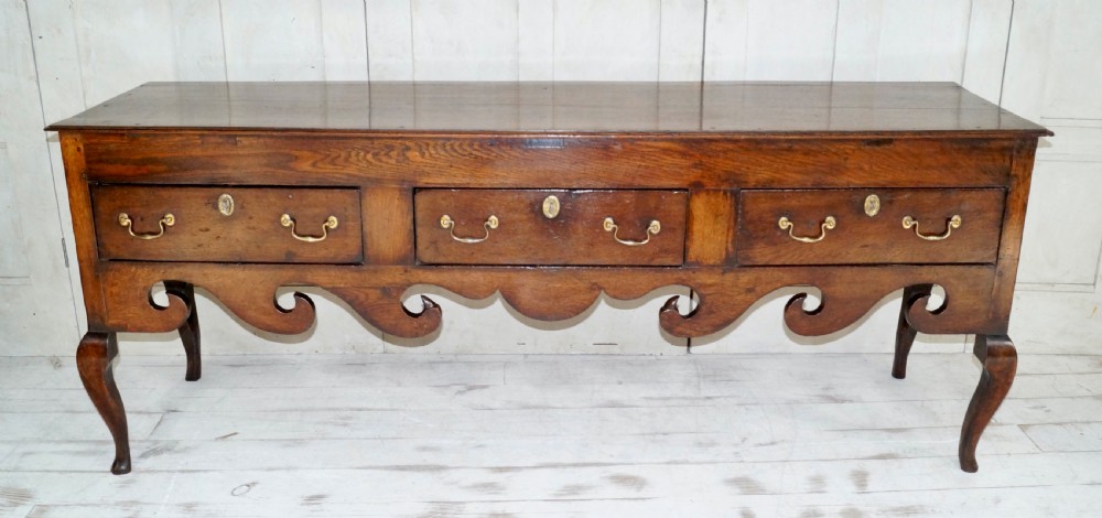An early Georgian dark oak dresser base featuring three drawers with brass handles, a distinctive scrolled apron, and cabriole legs. View XI
