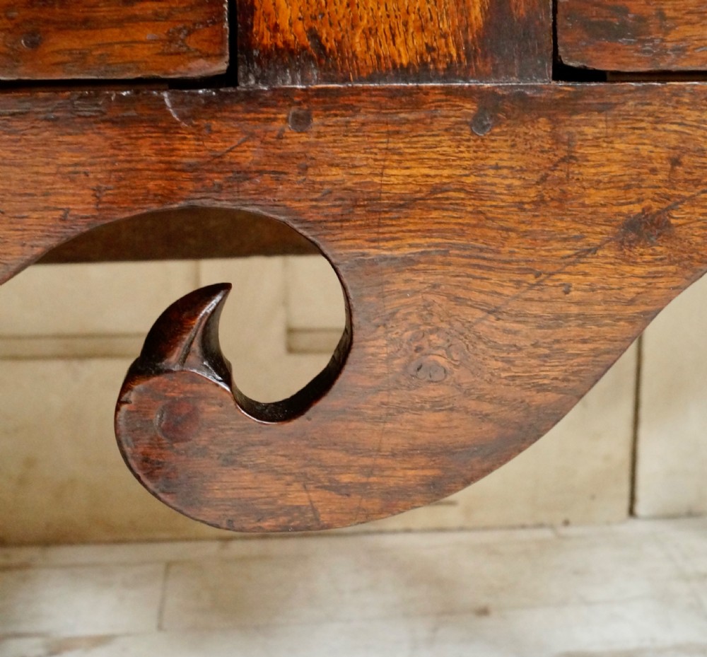 An early Georgian dark oak dresser base featuring three drawers with brass handles, a distinctive scrolled apron, and cabriole legs. View XII