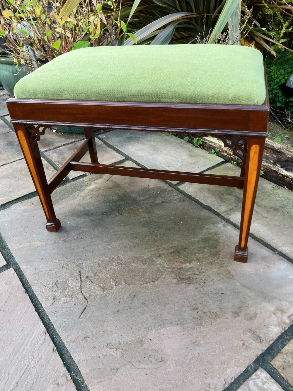 An Edwardian mahogany rectangular stool with light wood inlay on tapered legs, corner fretwork spandrels, an H-stretcher, and a green upholstered seat.
