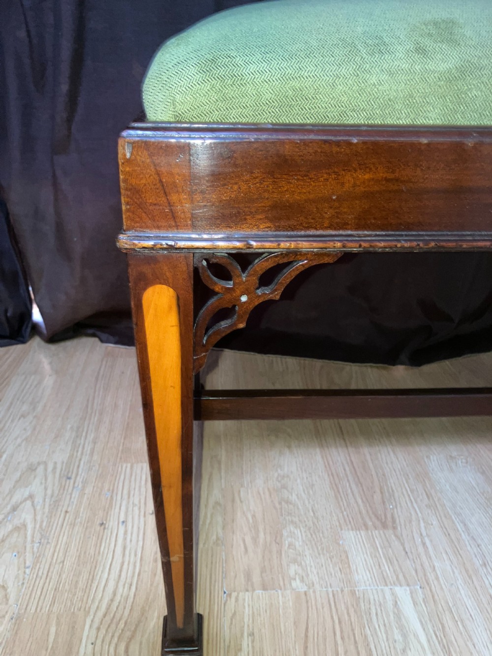 An Edwardian mahogany rectangular stool with light wood inlay on tapered legs, corner fretwork spandrels, an H-stretcher, and a green upholstered seat. View II