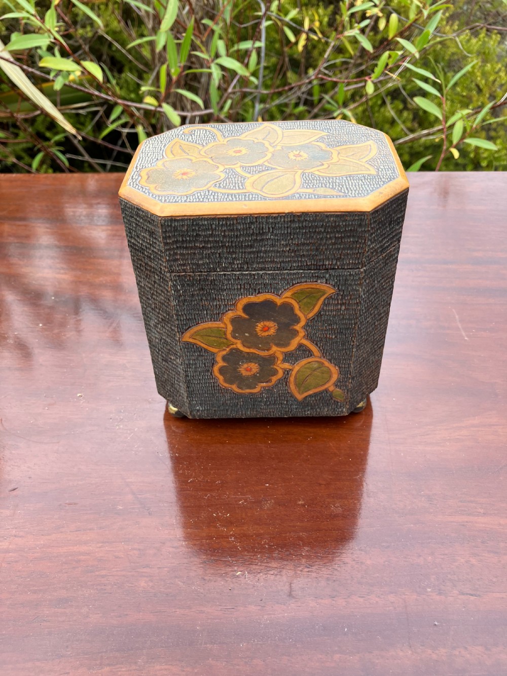 An octagonal wooden tea caddy featuring a textured stippled dark brown body, small brass feet, and painted yellow and red floral designs on the lid and front panel.