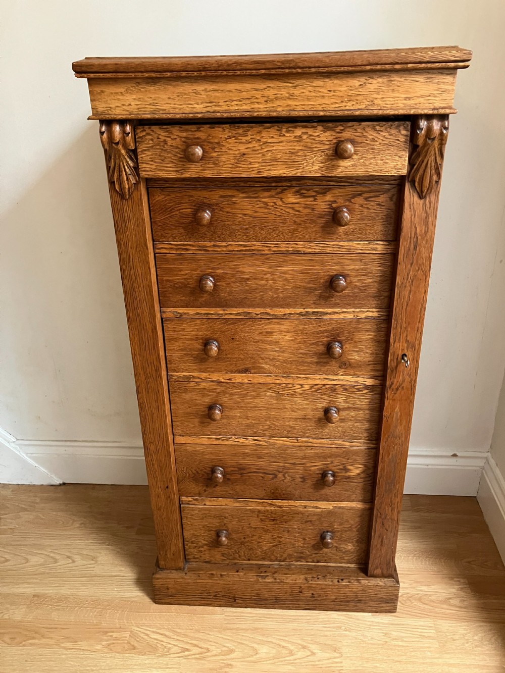 A tall, narrow Victorian oak Wellington chest with seven drawers, turned wooden knobs, carved upper corbels, and a side locking pilaster.
