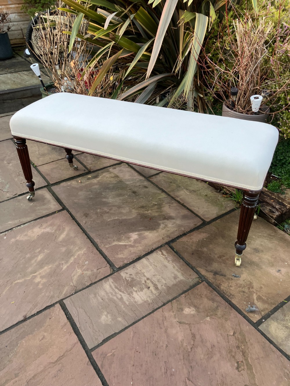 A long rectangular mahogany bench featuring cream leather upholstery supported by four reeded turned legs with brass castors.