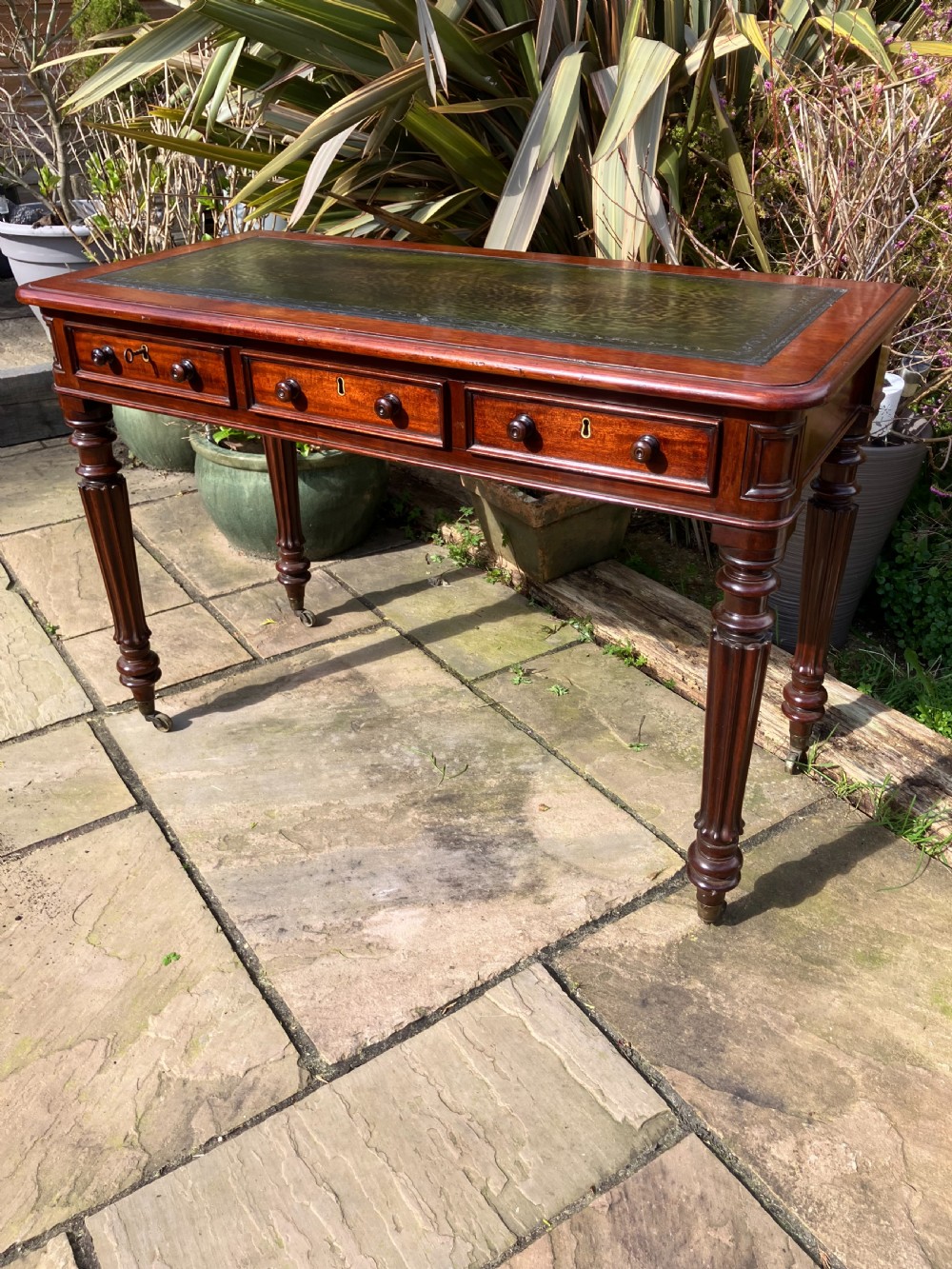 A Victorian mahogany writing table featuring a dark green leather inset top, three frieze drawers with wooden knobs, and turned fluted legs on castors.